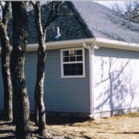 Light gray detached garage with two white doors and a single window, surrounded by leafless trees in winter.