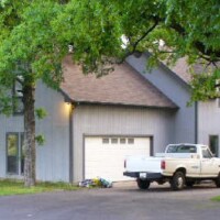 A white pickup truck is parked in front of a gray house with a double garage, surrounded by trees.