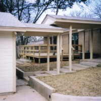 A covered concrete walkway connects a house to a detached garage, with a wooden ramp and deck in the background.