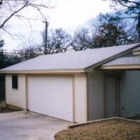 Detached two-car garage with white door and beige siding, surrounded by trees and a concrete driveway.
