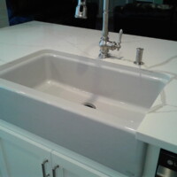 White farmhouse kitchen sink with stainless steel faucet set in a marble countertop on a white cabinet base.