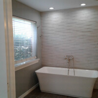 Modern bathroom with a white freestanding tub, wall-mounted faucet, tiled accent wall, and window with blinds.