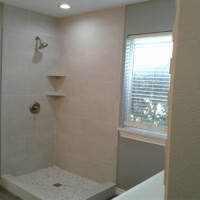Walk-in shower with beige tiles, two corner shelves, and a window with blinds next to the shower area.