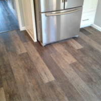 Stainless steel refrigerator on a kitchen floor with wood-look vinyl plank flooring in natural tones.