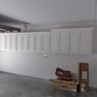 White wall cabinets are mounted in a garage above stacked cardboard boxes on a bare concrete floor.