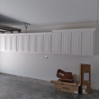 White wall cabinets are mounted in a garage above stacked cardboard boxes on a bare concrete floor.