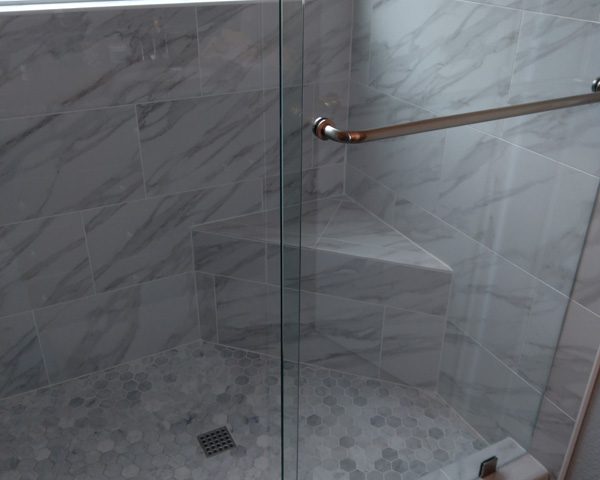 Glass-enclosed shower with gray marble tile, corner bench, and window above; toiletries on a built-in shelf.