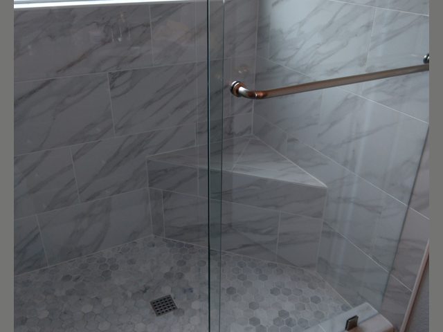 Glass-enclosed shower with gray marble tile, corner bench, and window above; toiletries on a built-in shelf.