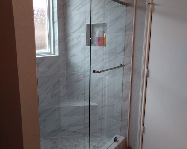 Modern shower with glass doors, marble tile walls, niche shelf with toiletries, a window, and a white door nearby.