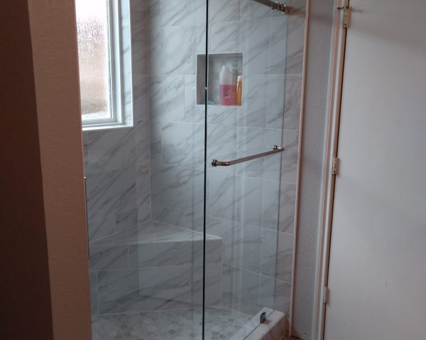 Modern shower with glass doors, marble tile walls, niche shelf with toiletries, a window, and a white door nearby.
