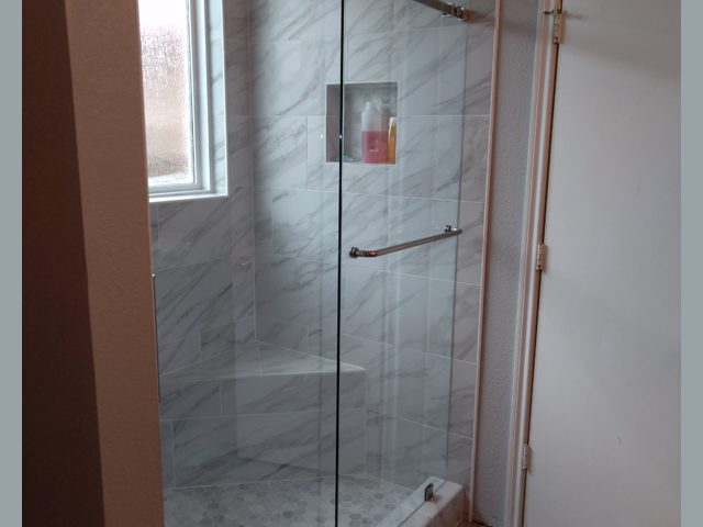 Modern shower with glass doors, marble tile walls, niche shelf with toiletries, a window, and a white door nearby.