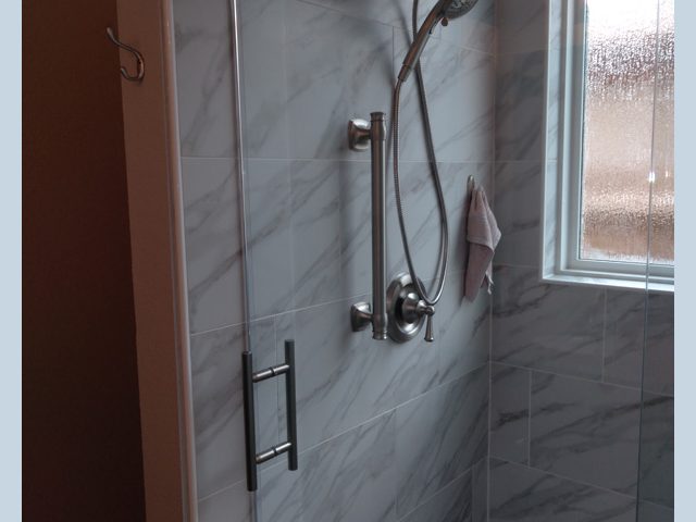 Tiled shower with a glass door, metal grab bar, handheld showerhead, and a towel hanging by a window.