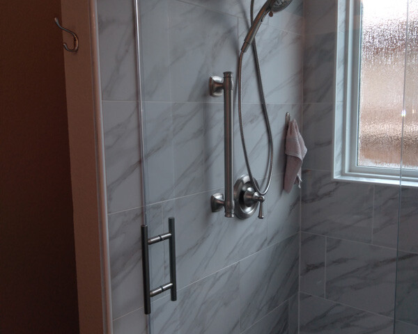 Tiled shower with a glass door, metal grab bar, handheld showerhead, and a towel hanging by a window.