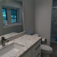 Modern bathroom with a marble countertop, white cabinets, toilet, and a large mirror under window light.
