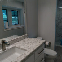 Modern bathroom with a marble countertop, white cabinets, toilet, and a large mirror under window light.