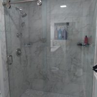 Glass shower enclosure with marble tile walls, pebble floor, and mounted showerhead; bottles on niche shelf.