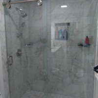 Glass shower enclosure with marble tile walls, pebble floor, and mounted showerhead; bottles on niche shelf.