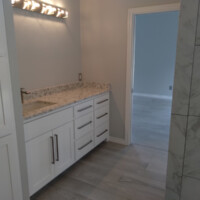 Modern bathroom with white cabinets, marble countertop, and tile flooring, adjacent to an empty room with wood flooring.