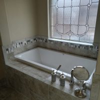 A built-in bathtub with decorative tile, a frosted window, and toiletries on the ledge in a bathroom.