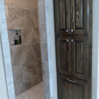 Tall wooden cabinet next to a tiled walk-in shower with built-in shelves and mosaic floor tiles.