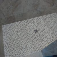 Shower floor with basketweave-patterned tiles and a central square drain, surrounded by large gray wall tiles.