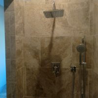 A tiled shower with a square overhead showerhead, a handheld shower, and a control panel; a toilet is visible in the adjacent room.