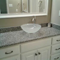 Bathroom vanity with granite countertop, vessel sink, chrome faucet, framed mirror, and light-colored cabinets.