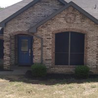 Front view of a brick house with a blue door, two large windows, and shrubs along the entrance walkway.