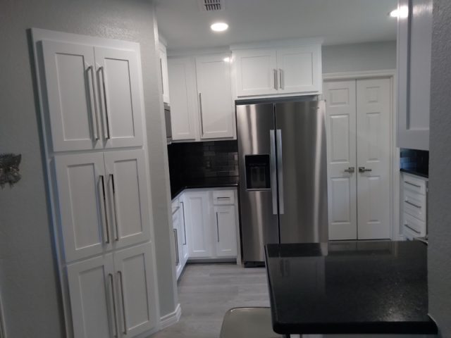 Modern kitchen with white cabinets, stainless steel refrigerator, black countertops, and light gray flooring.