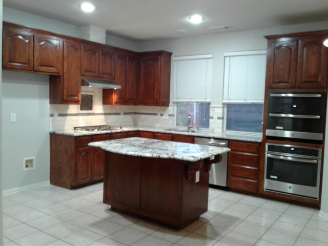Kitchen with dark wood cabinets, a marble island, double ovens, gas stove, tile floor, and two windows.