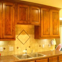 Wooden kitchen cabinets above a tiled backsplash with a double sink and countertop beneath.