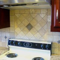 4 inch tumbled marble stone tile with bronze medallions Electric stove with four burners, control panel, and tiled backsplash beneath a range hood and wooden cabinets.