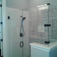 Glass shower enclosure with black fixtures and patterned tile wall, adjacent to a bedroom with a white bed.