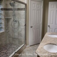Modern bathroom with a glass-enclosed shower, double sinks, granite countertop, and two white doors.