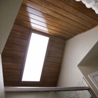 Wood-paneled bathroom ceiling with a rectangular skylight above a glass shower enclosure and vanity lights.